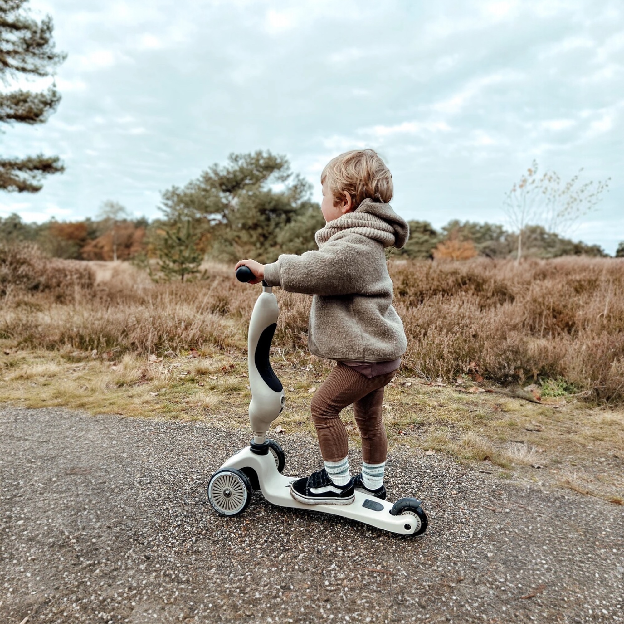 Väikelaps halli Scoot and Ride Highwaykick 1 tõukerattaga looduse keskel, seismas ja nautimas sõitu.