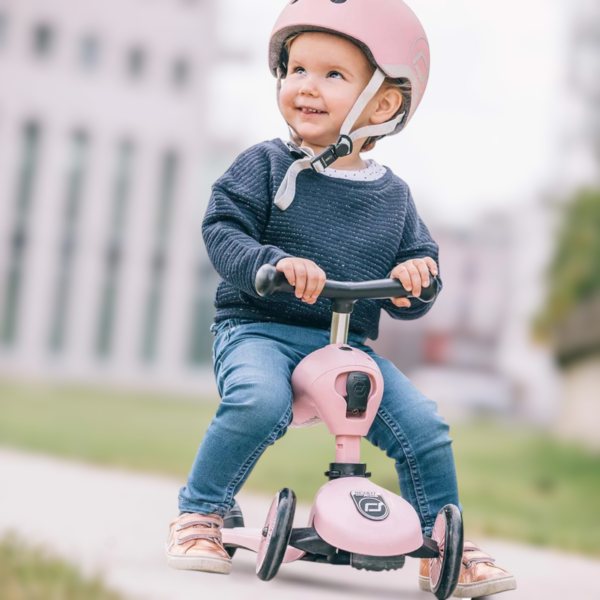 Väikelaps roosa Scoot and Ride Highwaykick 1 tõukerattaga, kandes vastavat kiivrit, naeratamas ja nautimas sõitu õues.